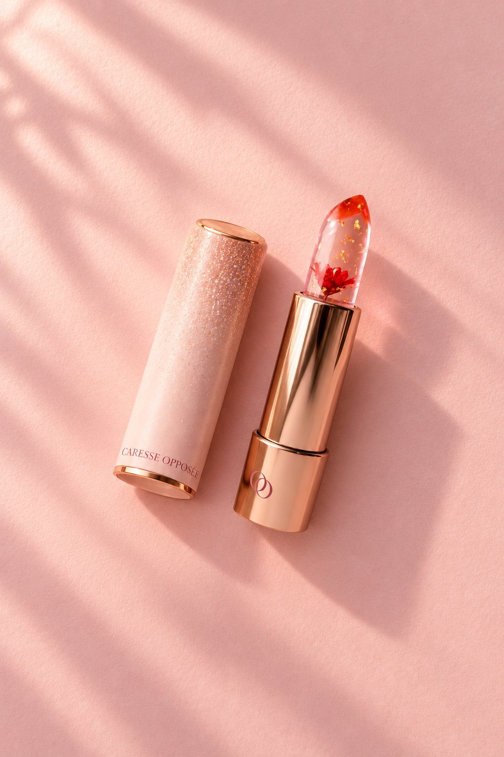 Baume à lèvres Caresse Opposée ouvert, avec sa fleur rouge visible dans le stick transparent et son capuchon rose pailleté posé à côté, sur fond rose.