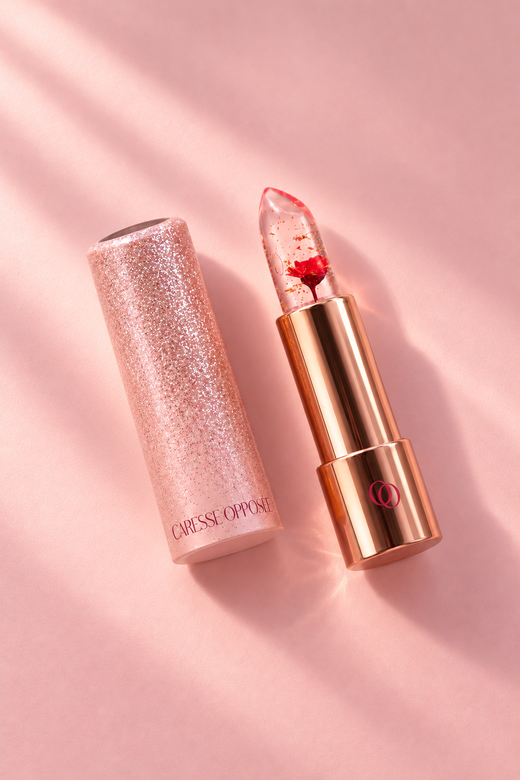 Baume à lèvres Caresse Opposée ouvert, avec sa fleur rouge visible dans le stick transparent et son capuchon rose pailleté posé à côté, sur fond rose.