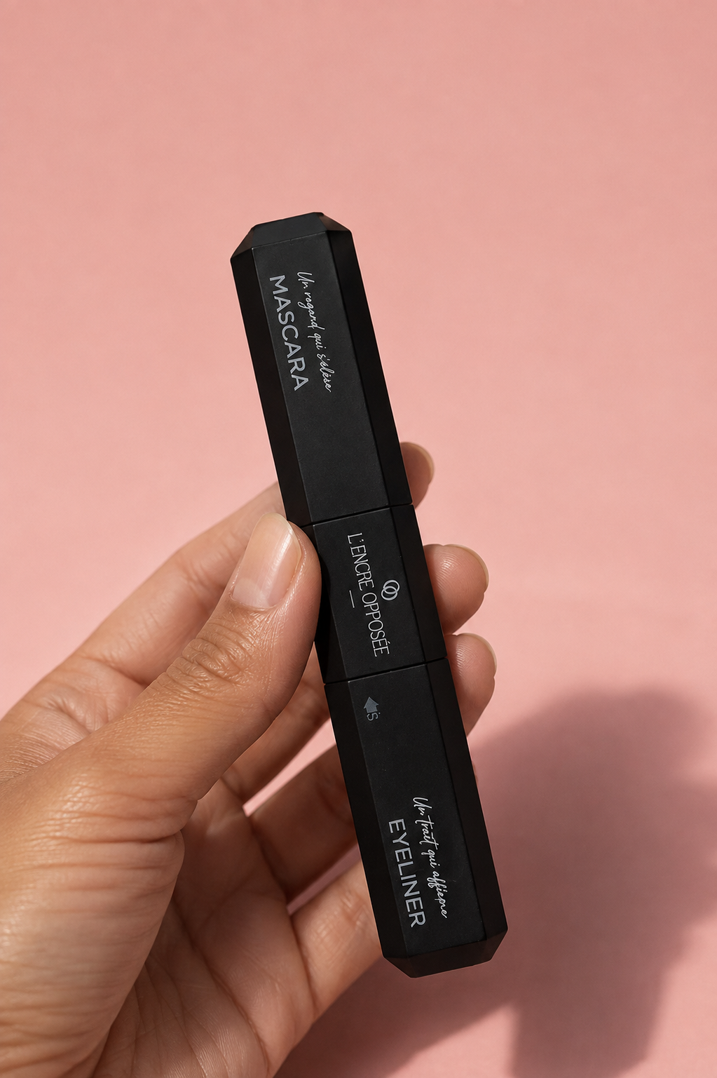 L’Encre Opposée tenue en main sur fond rose, mascara et eyeliner réunis dans un seul produit.
