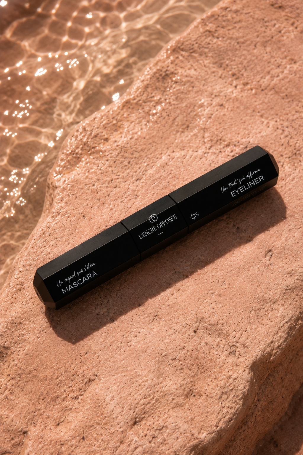 Mascara et eyeliner 2-en-1 L’Encre Opposée posé sur une pierre rose au bord de l’eau.