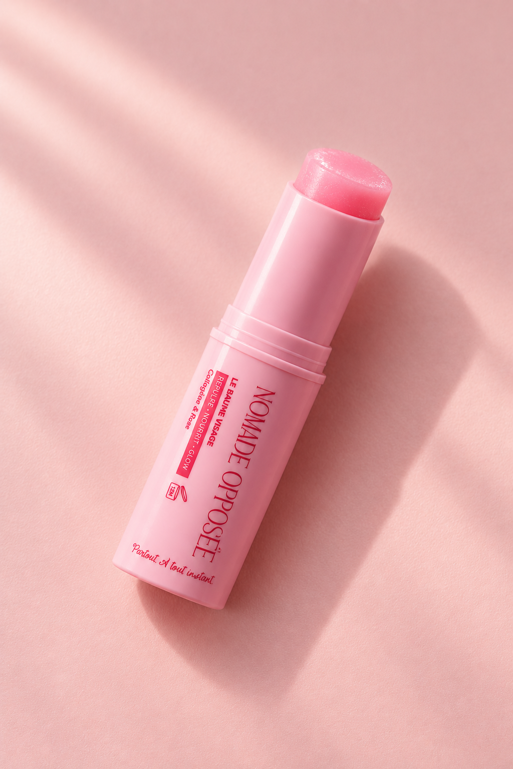 Baume visage Nomade Opposée ouvert et allongé sur fond rose, avec sa texture rosée visible, un reflet lumineux sur le tube et une ombre douce.